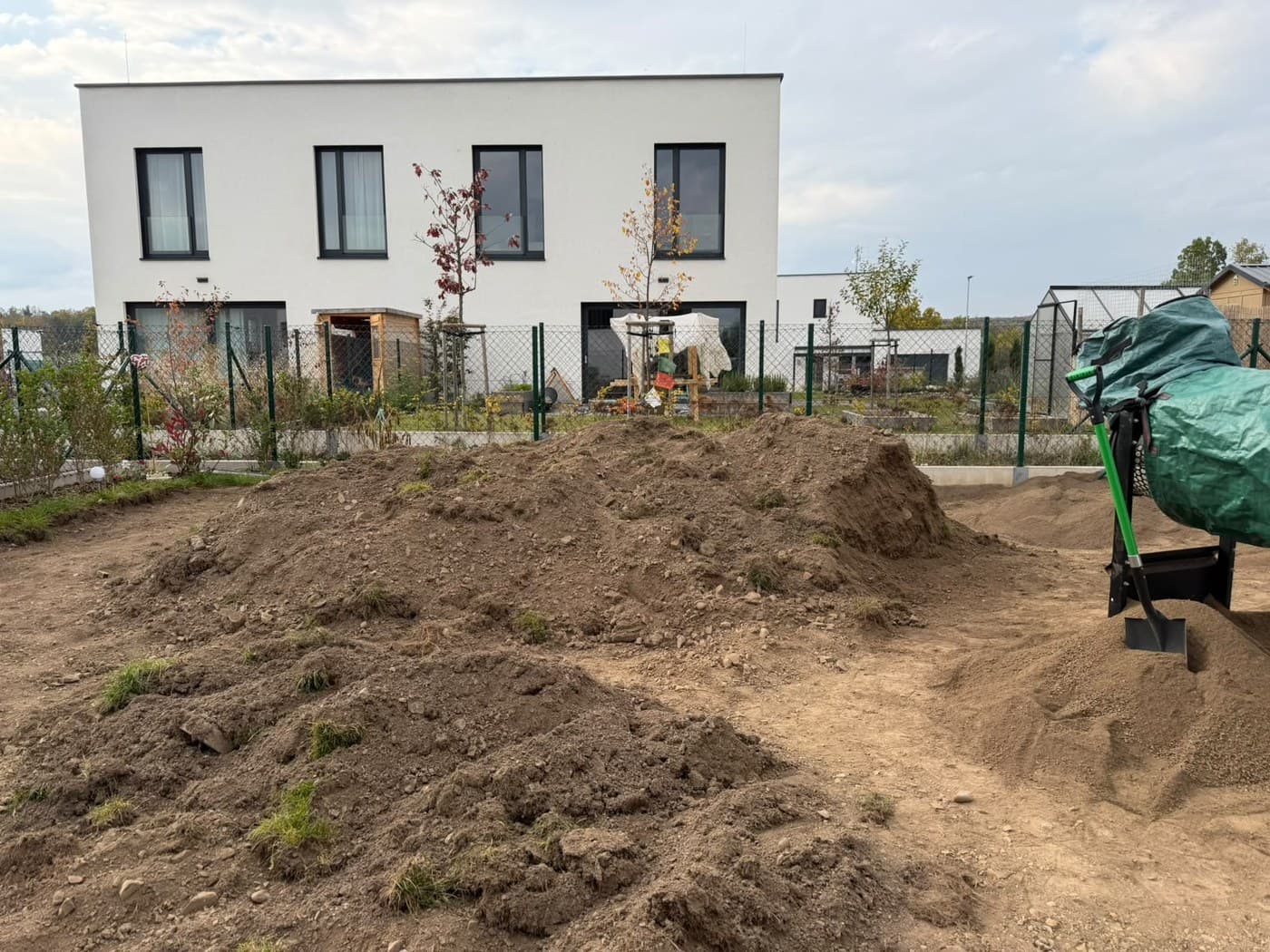 Zahrada u novostavby s hromadami navezené zeminy, lopatou a zakrytým materiálem v rohu