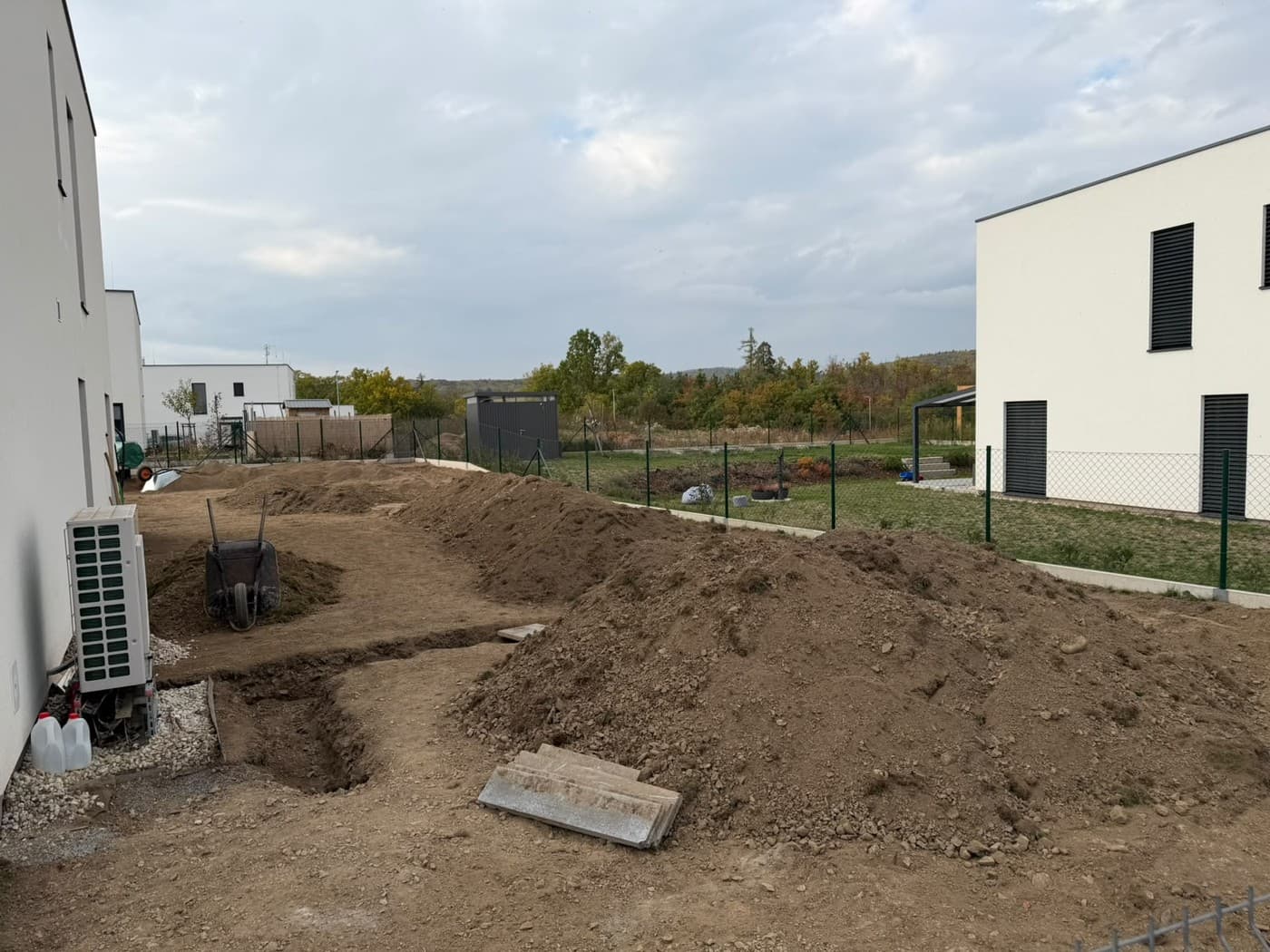 Pruh zahrady podél domu během prací, hromady zeminy, výkop a kolečko u venkovní jednotky